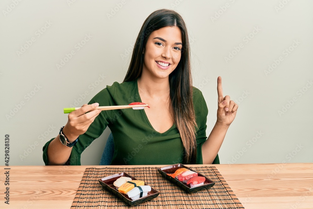 Sticker Young brunette woman eating sushi sitting on the table smiling with an idea or question pointing finger with happy face, number one