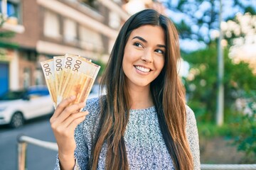 Young beautiful hispanic girl smiling happy holding norwegian krone banknotes at the city.