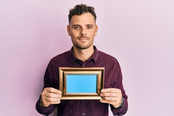 Young hispanic man holding empty frame relaxed with serious expression on face. simple and natural looking at the camera.