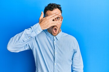 Young hispanic man wearing casual clothes and glasses peeking in shock covering face and eyes with hand, looking through fingers with embarrassed expression.