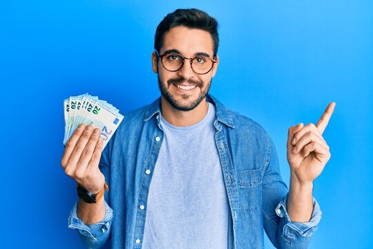 Young hispanic man holding bunch of 20 euro banknotes smiling happy pointing with hand and finger to the side