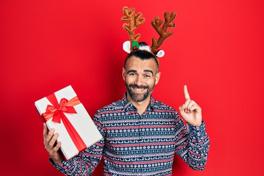 Young hispanic man wearing deer christmas hat holding gift smiling with an idea or question pointing finger with happy face, number one
