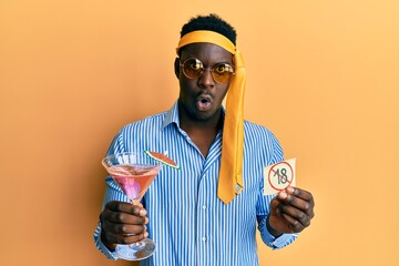 Handsome black man drunk wearing tie over head drinking cocktail holding under 18 sign in shock...