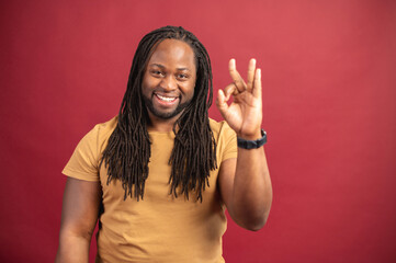 Charismatic dark skinned guy with a long dreadlocks showing OK gesture isolated on red background,...