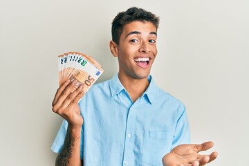 Young handsome african american man holding bunch of 50 euro banknotes celebrating achievement with happy smile and winner expression with raised hand