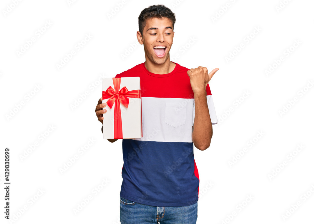 Wall mural Young handsome african american man holding gift pointing thumb up to the side smiling happy with open mouth