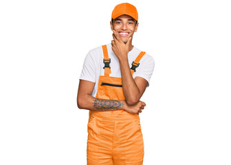Young handsome african american man wearing handyman uniform looking confident at the camera smiling with crossed arms and hand raised on chin. thinking positive.
