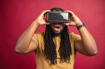 Black amazed man with long dreadlocks dressed in casual clothes, standing isolated over red background with virtual reality headset, glasses in an empty room, traveling online, trying new experience