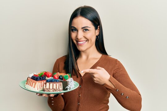 Young brunette woman holding plate with cake slices smiling happy pointing with hand and finger
