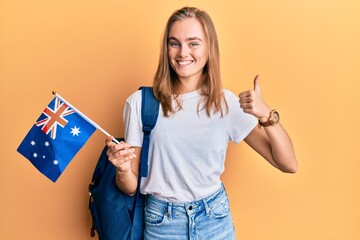 Beautiful blonde woman exchange student holding australia flag smiling happy and positive, thumb up doing excellent and approval sign