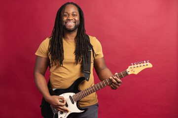 Smiling energetic and enthusiastic young African American male musician with dreadlocks playing electric guitar isolated on red wall, giving virtual classes, streaming performing online