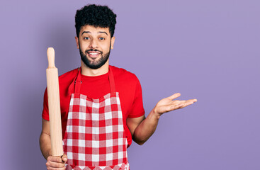 Young arab man with beard wearing professional baker apron holding kneading roll celebrating achievement with happy smile and winner expression with raised hand