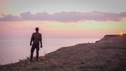 Looking on young biker in leather suit stay on a precipice above sea looking far on majestic sunset with rose clouds. 