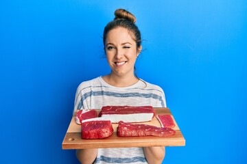 Young blonde woman holding board with raw meat winking looking at the camera with sexy expression,...
