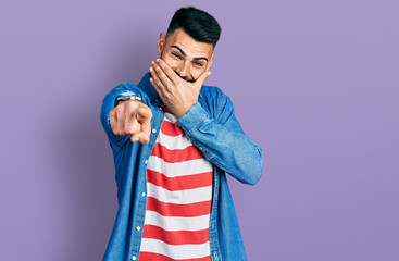 Young hispanic man with beard wearing casual denim jacket laughing at you, pointing finger to the camera with hand over mouth, shame expression