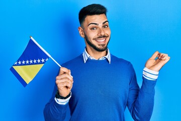 Young hispanic man with beard holding bosnia herzegovina flag screaming proud, celebrating victory and success very excited with raised arm