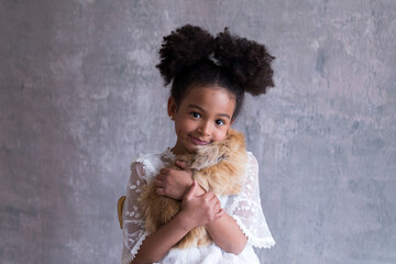 Medium shot of adorable mixed race little girl sitting in a lacy white dress holding a cute long-haired caramel rabbit tight to her chest