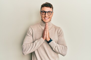 Hispanic young man wearing casual turtleneck sweater praying with hands together asking for forgiveness smiling confident.