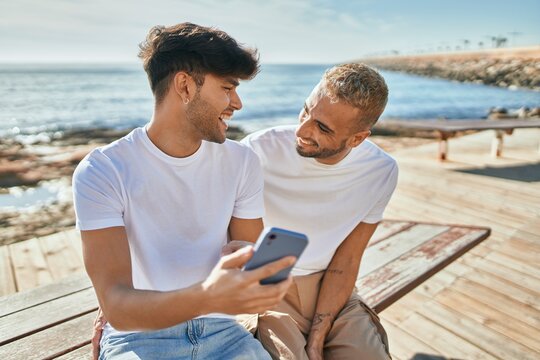 Young gay couple smiling happy using smartphone at the beach promenade.
