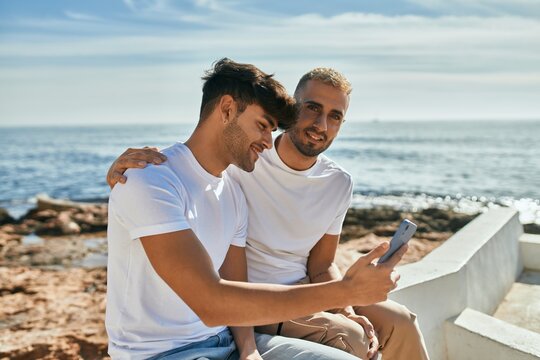Young gay couple smiling happy using smartphone at the beach.