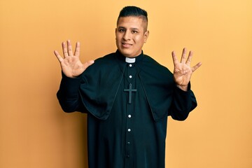 Young latin priest man standing over yellow background showing and pointing up with fingers number...