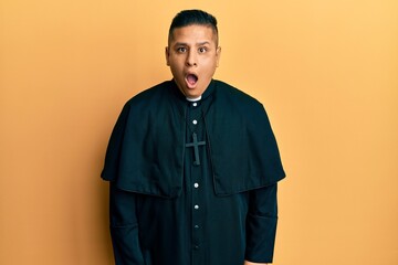 Young latin priest man standing over yellow background afraid and shocked with surprise and amazed expression, fear and excited face.