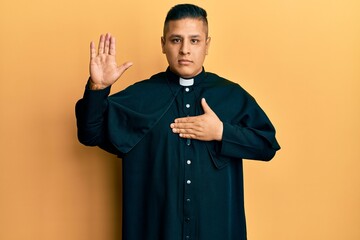 Young latin priest man standing over yellow background swearing with hand on chest and open palm,...