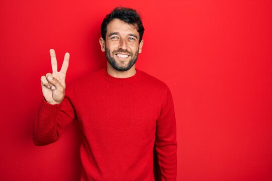 Handsome man with beard wearing casual red sweater smiling looking to the camera showing fingers doing victory sign. number two.