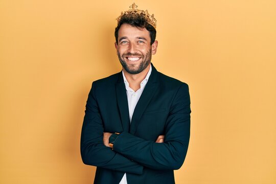 Handsome man with beard wearing business suit with arms crossed gesture and king crown smiling with a happy and cool smile on face. showing teeth.
