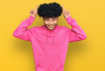 Young african american man with afro hair wearing casual pink sweatshirt posing funny and crazy with fingers on head as bunny ears, smiling cheerful