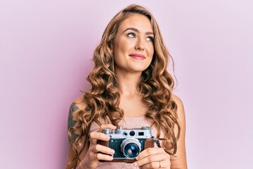 Young blonde girl holding vintage camera smiling looking to the side and staring away thinking.