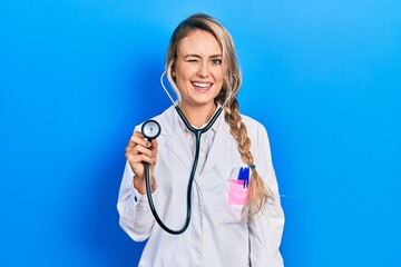Beautiful young blonde doctor woman holding stethoscope winking looking at the camera with sexy...