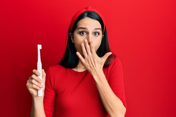Young hispanic woman holding electric toothbrush covering mouth with hand, shocked and afraid for...