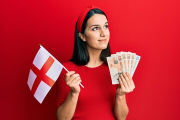 Young hispanic woman holding england flag and pounds banknotes smiling looking to the side and staring away thinking.