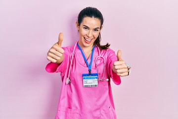 Young brunette woman wearing doctor uniform and stethoscope approving doing positive gesture with hand, thumbs up smiling and happy for success. winner gesture.