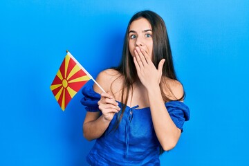 Young brunette teenager holding macedonian flag covering mouth with hand, shocked and afraid for...