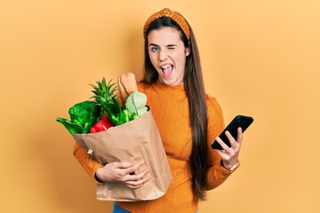 Young brunette teenager holding bag of groceries using smartphone winking looking at the camera with sexy expression, cheerful and happy face.