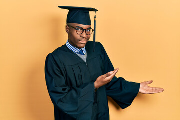 Young african american man wearing graduation cap and ceremony robe inviting to enter smiling...