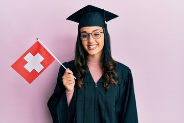 Young hispanic woman wearing graduation uniform holding switzerland flag looking positive and happy standing and smiling with a confident smile showing teeth