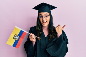 Young hispanic woman wearing graduation uniform holding ecuador flag pointing thumb up to the side smiling happy with open mouth