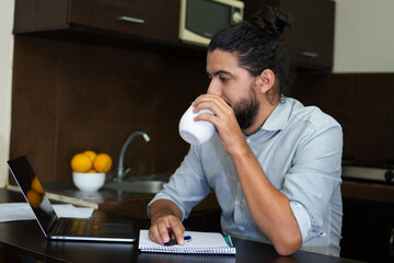 young adult having a drink sitting relaxed in the kitchen while looking at the laptop next to a note pad, working at home.