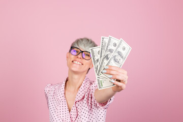 Stylish woman in dress and glasses on pink background pulling money to you, fan of 100 dollars  happy  positive cheerful