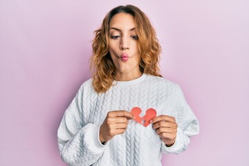 Young caucasian woman holding broken heart paper shape making fish face with mouth and squinting...