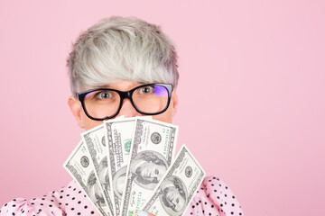 Stylish woman in dress and glasses on pink background holding money fan of 100 dollars cover face close portrait