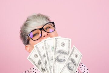 Stylish woman in dress and glasses on pink background holding money fan of 100 dollars cover face close portrait