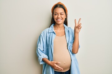 Beautiful hispanic woman expecting a baby, touching pregnant belly smiling with happy face winking...