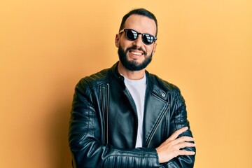 Young man with beard wearing black leather jacket and sunglasses happy face smiling with crossed...