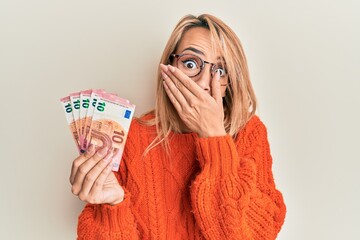 Beautiful blonde woman holding 10 euro banknotes covering mouth with hand, shocked and afraid for...