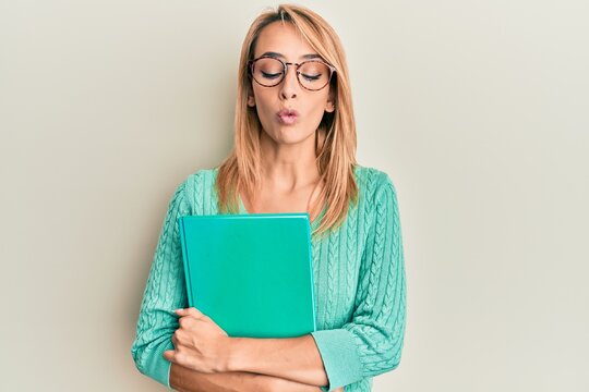 Beautiful blonde woman holding book wearing glasses making fish face with mouth and squinting eyes, crazy and comical.