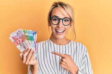 Beautiful blonde woman holding hong kong dollars banknotes smiling happy pointing with hand and finger
